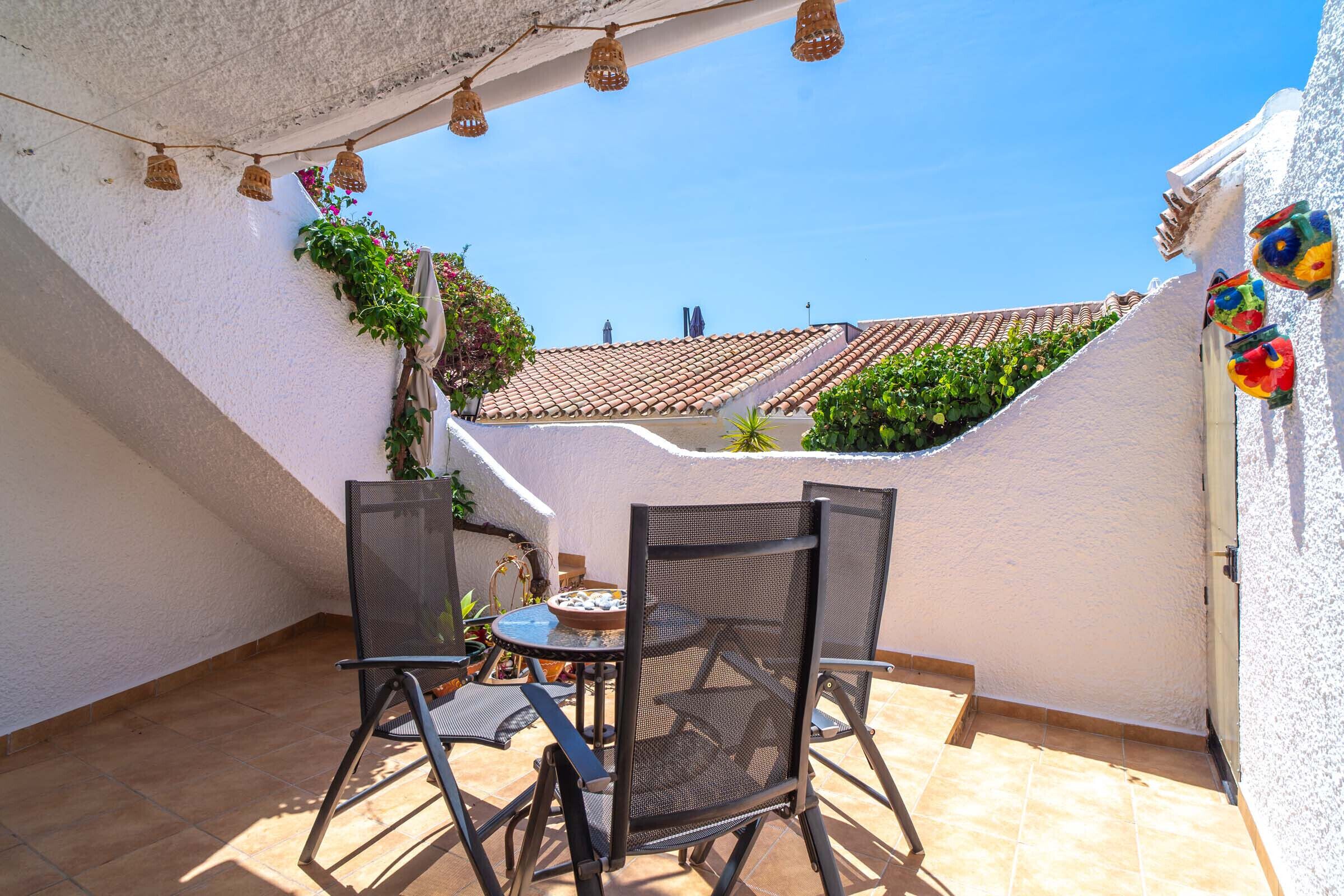 A patio with a table and chairs, a tiled floor, and a white wall with a tiled roof visible in the background.