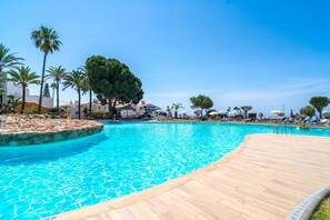 A swimming pool with a wooden deck, surrounded by palm trees and lounge chairs.