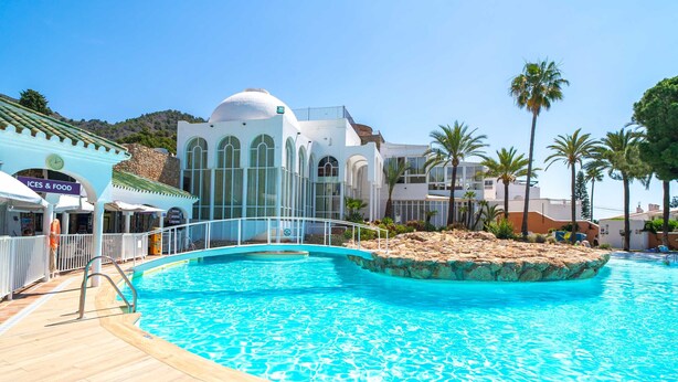 Una piscina con cubierta de madera, un edificio blanco con ventanas arqueadas y palmeras.