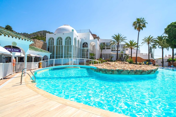 A swimming pool with a wooden deck, a white building with arched windows, and palm trees.