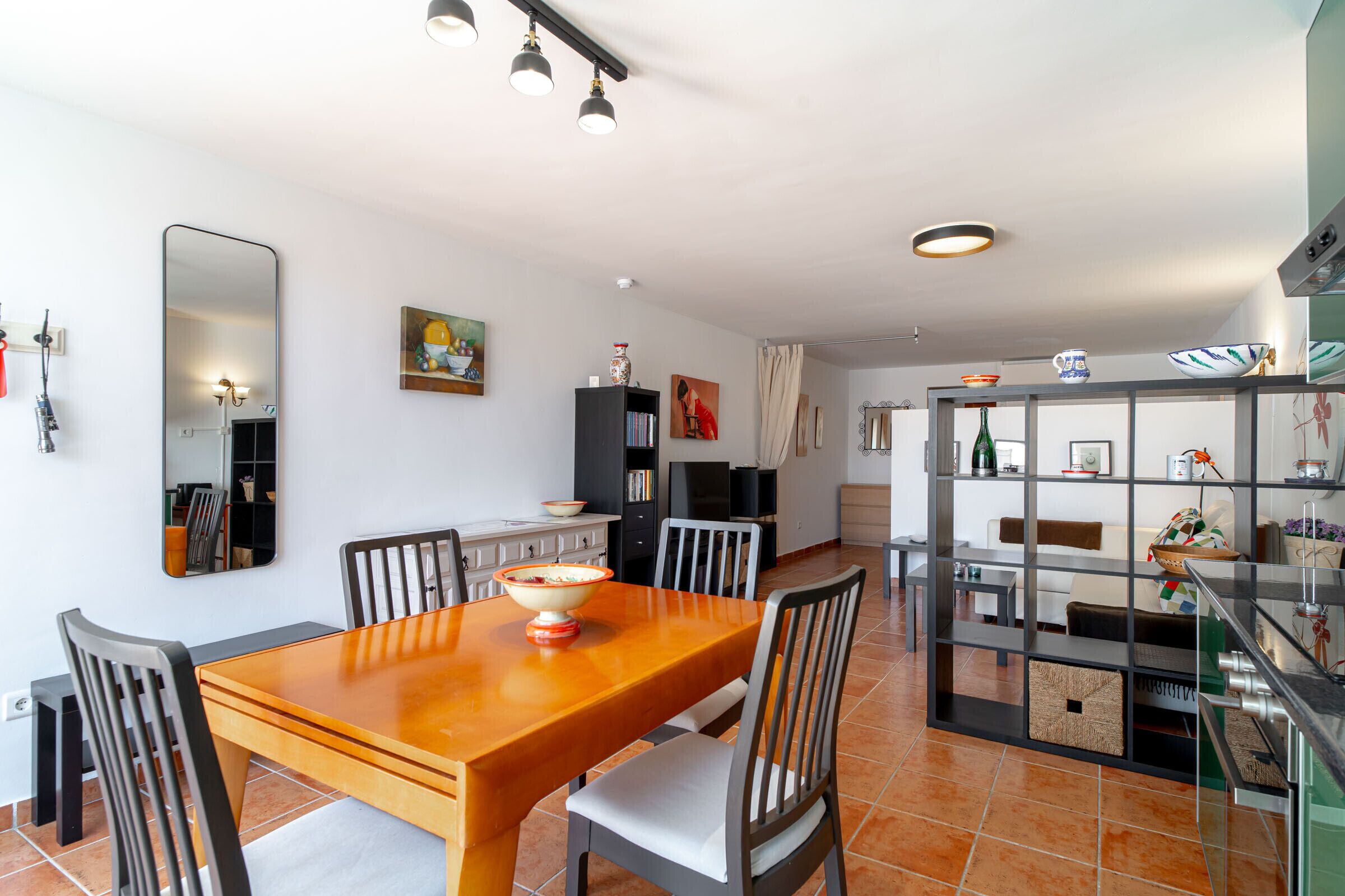 A dining area with a wooden table and chairs, a kitchen area with cabinets and a refrigerator, and a living space with a sofa and a TV.