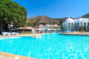 A swimming pool with clear blue water, surrounded by lounge chairs and umbrellas, with a white building and a hill in the background.
