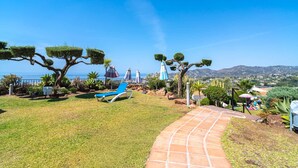 Un jardín con un sendero, una silla de descanso azul y vista al mar.