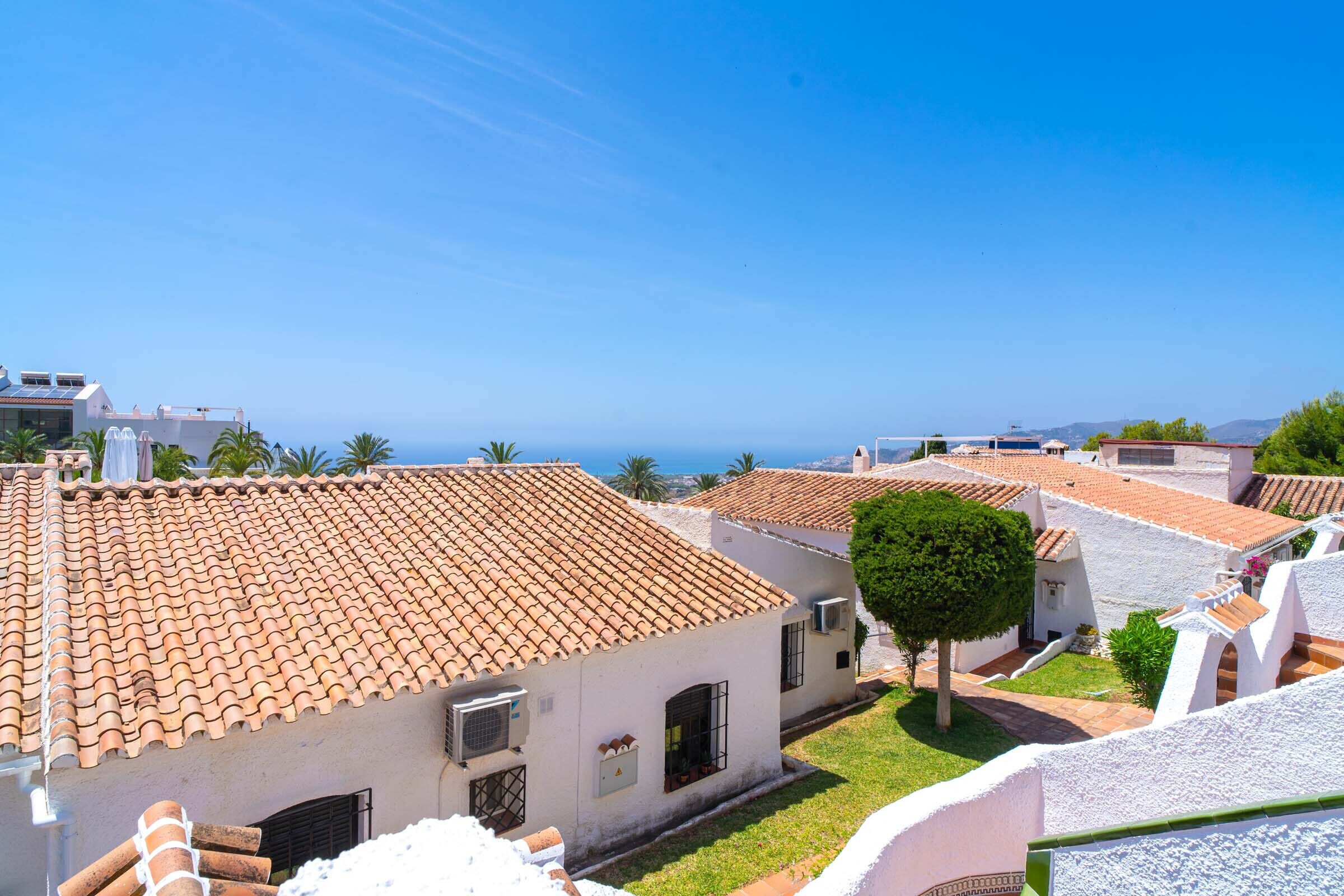 A coastal residential area with terracotta-tiled roofs, white walls, and a clear view of the sea.