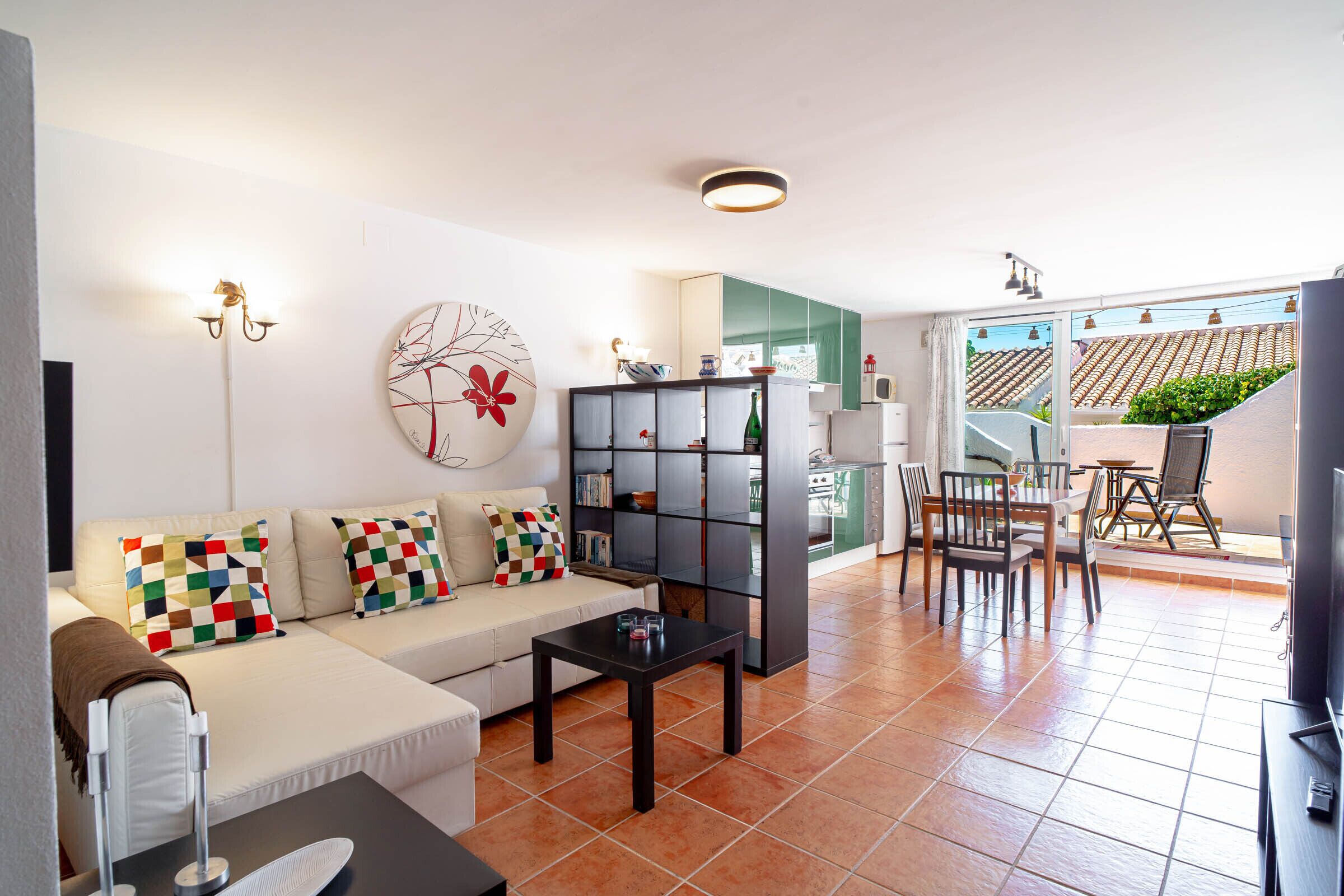 A modern living room with a sofa, coffee table, and a bookshelf. There is a dining area with chairs and a table. The room has a large window and a view of the outside.