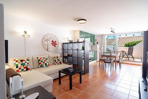 A modern living room with a sofa, coffee table, and a bookshelf. There is a dining area with chairs and a table. The room has a large window and a view of the outside.
