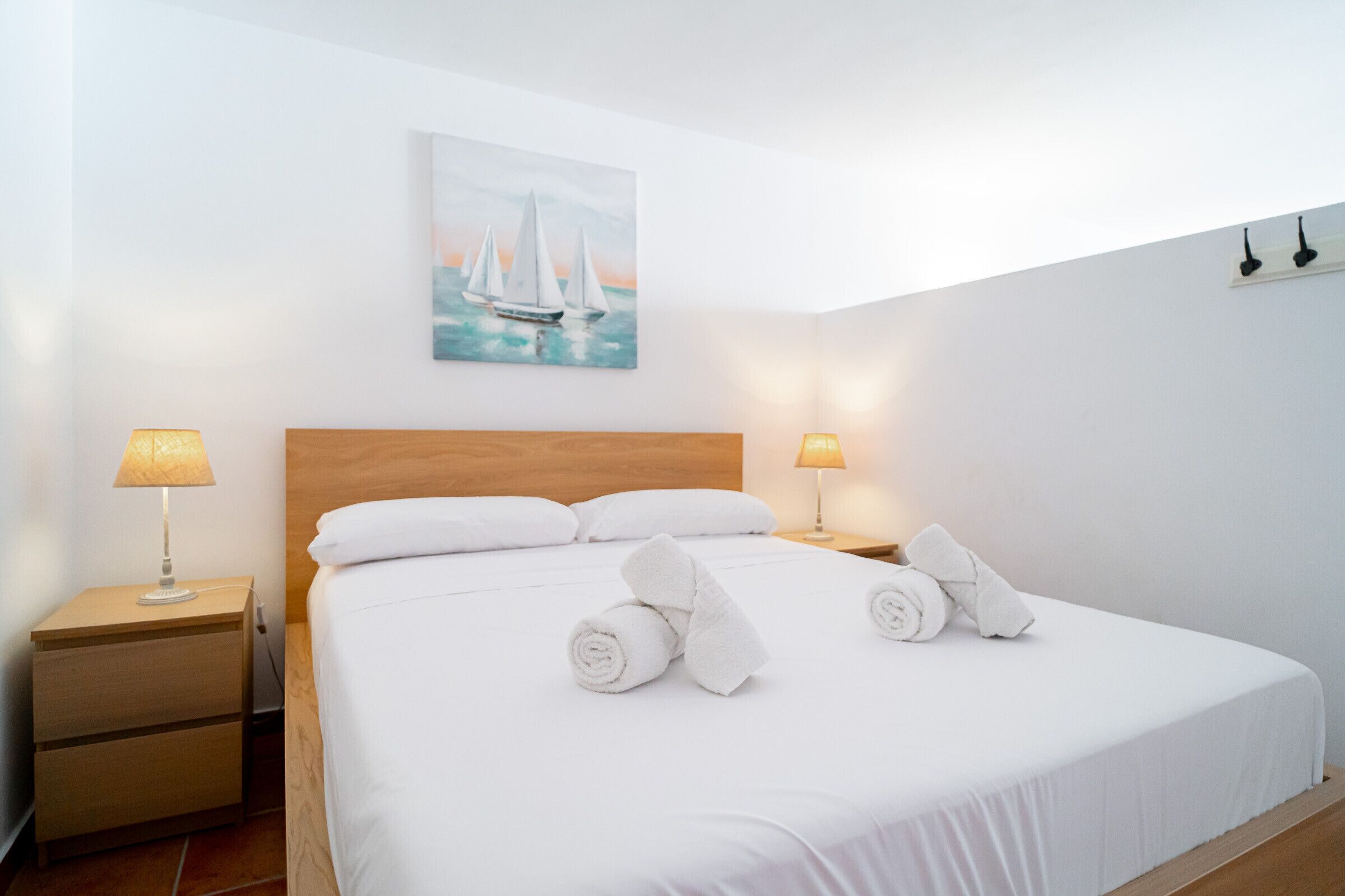 Een net opgemaakt bed met witte lakens, twee bedlampen, een houten hoofdbord en een schilderij van zeilboten aan de muur.