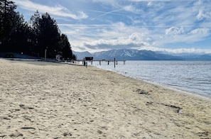 A sandy beach with a pier extending into calm waters, surrounded by trees and mountains in the background.