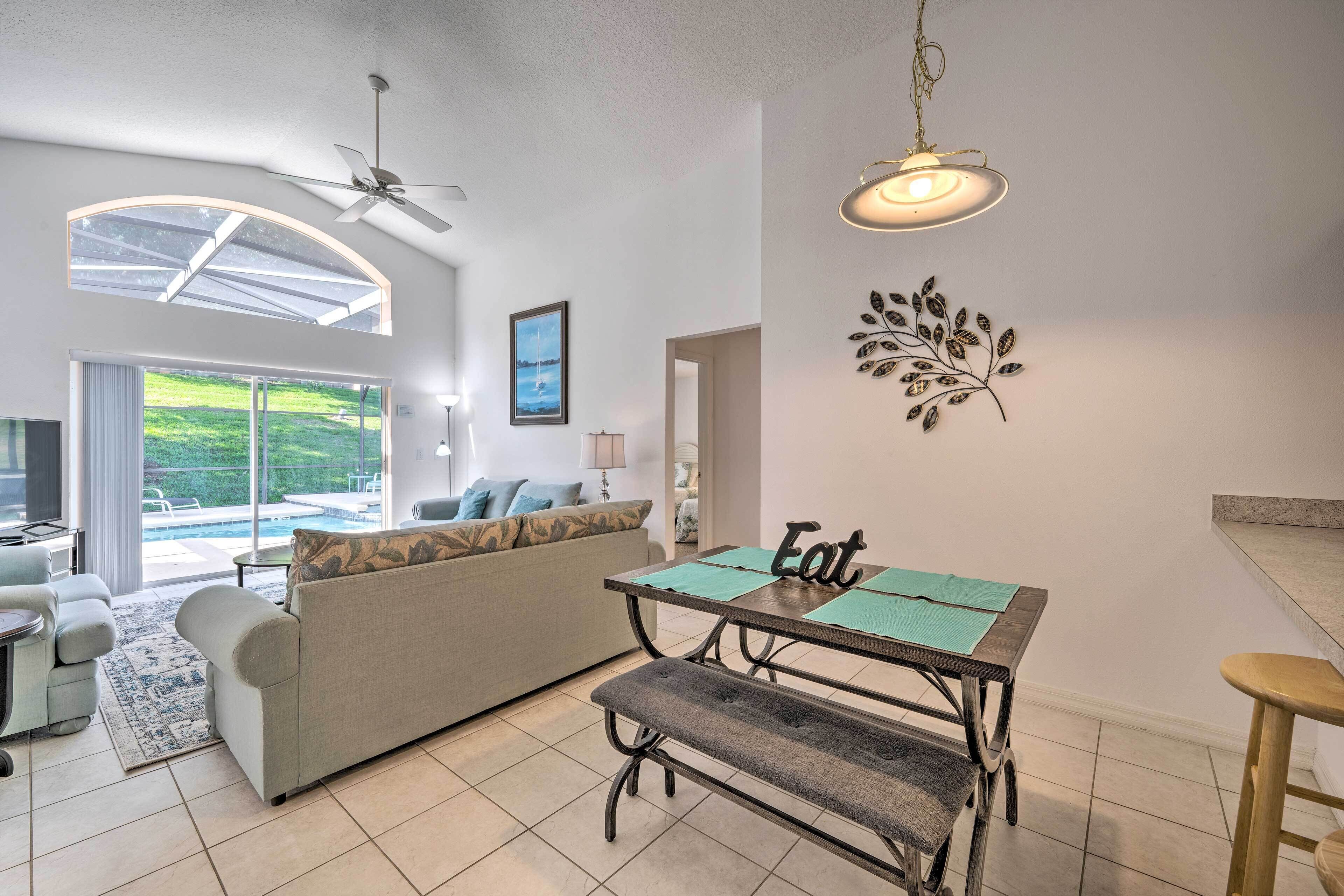 Uma sala de estar moderna com um sofá, uma mesa de jantar com um banco, um ventilador de teto e vista para uma piscina através de grandes janelas.