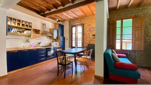 Une cuisine avec des meubles de rangement bleus, une table à manger et des chaises. On y trouve un mur en pierre et des poutres en bois au plafond.