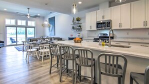 A modern kitchen with white cabinets, a large island with bar stools, a ceiling fan, and a dining area with a large window. - Family-Friendly Home: 4 Mi to Bethany Beach! (Millville)
