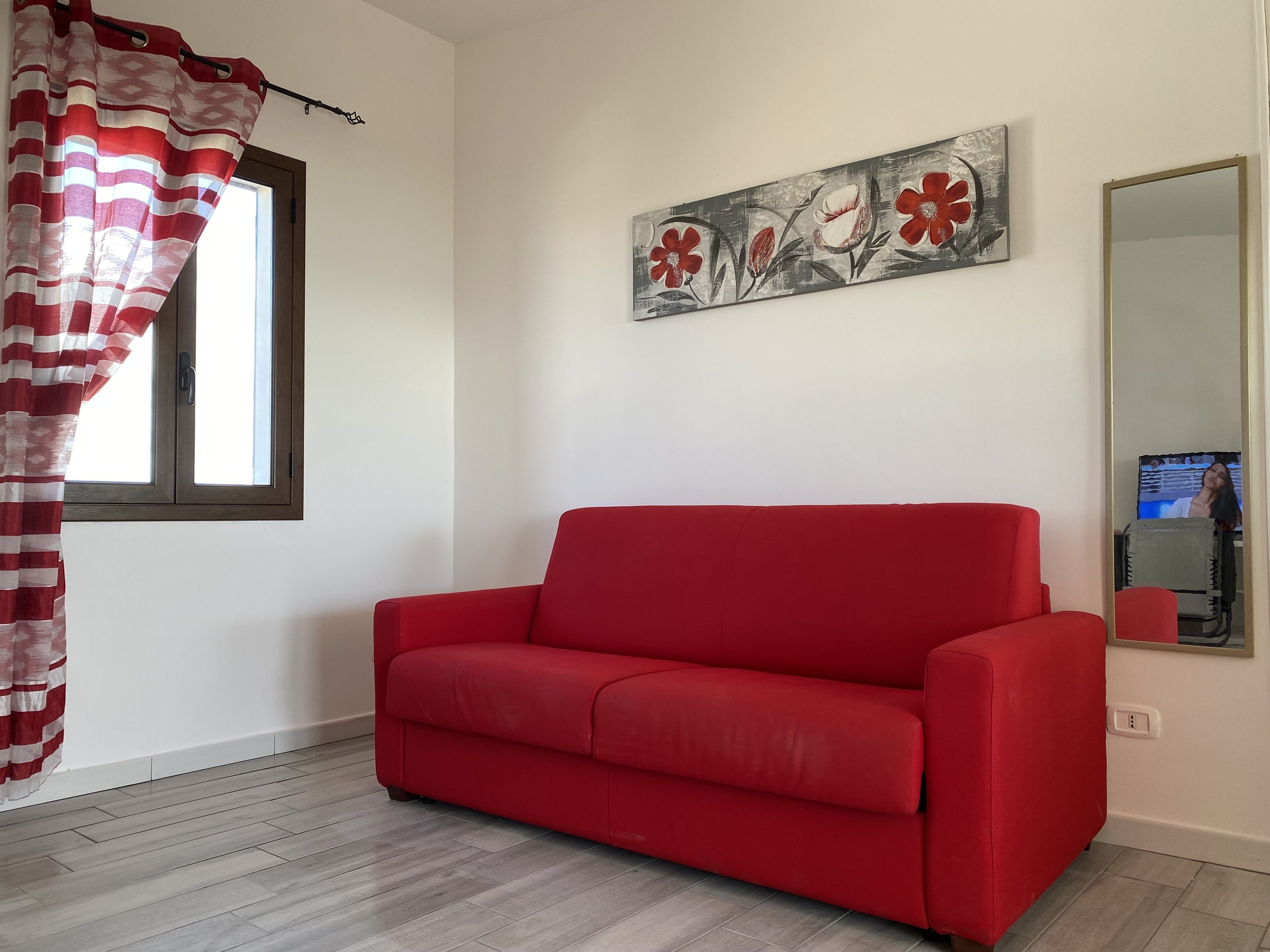 A red sofa in a room with a window, a painting on the wall, and a TV on a stand.