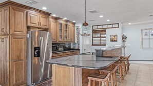 A kitchen with wooden cabinets, a stainless steel refrigerator, a granite countertop, and bar stools. - Gulf Access, Minutes to the River, Kayaks & Dog Friendly - Aqua Escape - Roelens Vacations (Cape Coral)