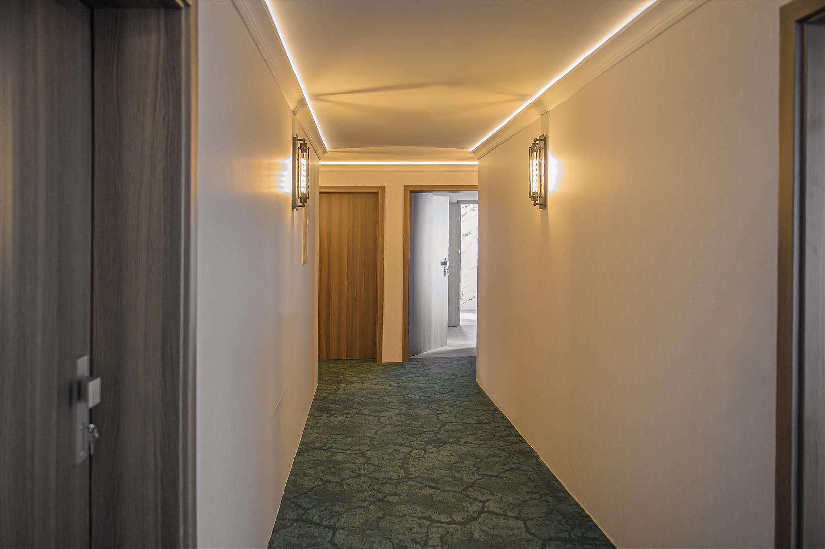 A hallway with a patterned carpet, wooden doors, and wall-mounted lighting fixtures.