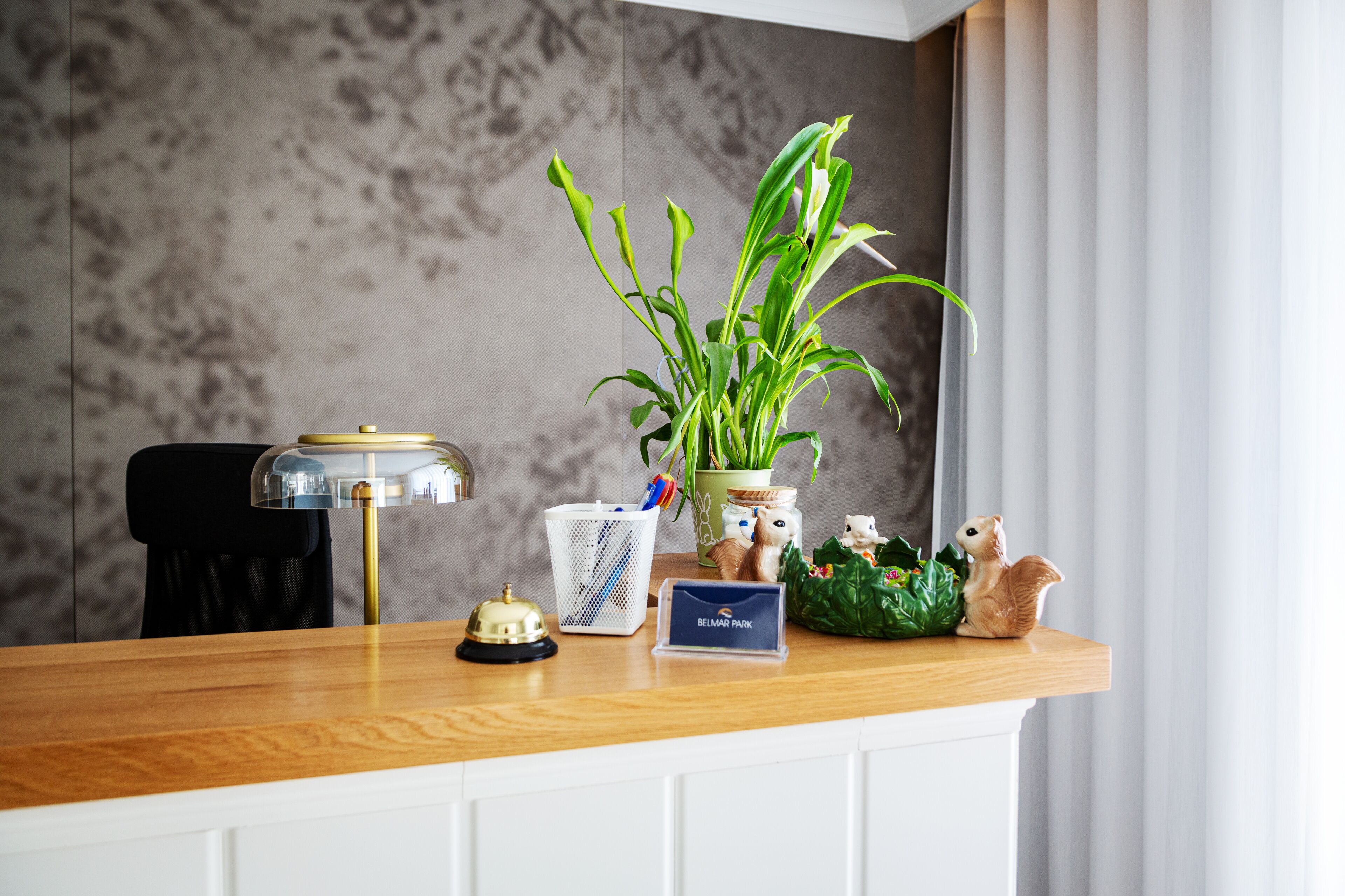 A reception desk with a wooden top, a brass bell, a sign reading "BELMAR PARK," a potted plant, a basket with pens, and a squirrel figurine.