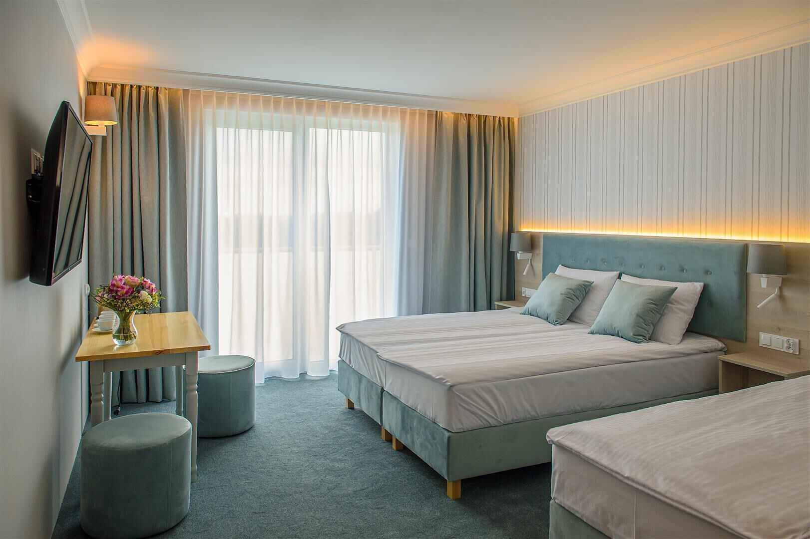 Une chambre d’hôtel avec un lit, des tables de chevet, une télévision, une lampe, une petite table ornée d’un vase de fleurs et une fenêtre avec des rideaux.