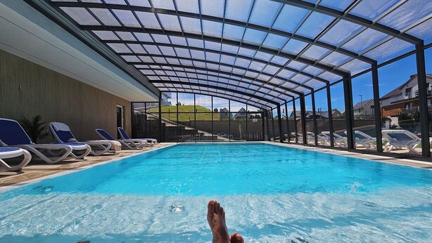 A swimming pool with a glass ceiling, lounge chairs, and a view of houses in the background. - Belmar Park (Wladyslawowo)