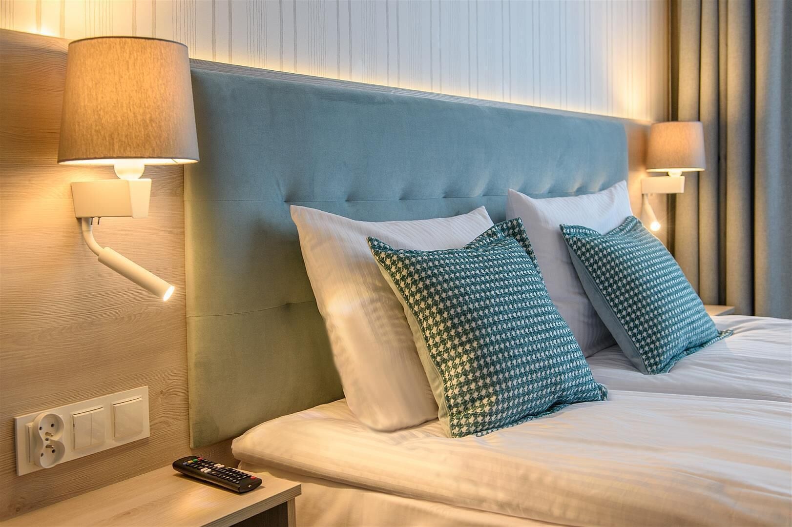 A hotel room with a tufted headboard, two bedside lamps, a remote control, and patterned pillows.