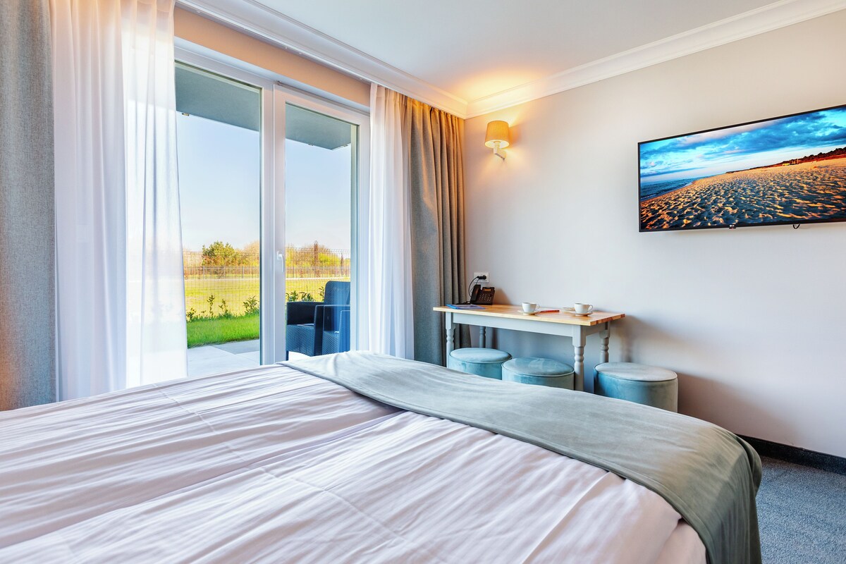 Habitación de hotel con una cama grande, un escritorio con dos sillas, vista a una playa a través de la ventana y un cuadro en la pared.