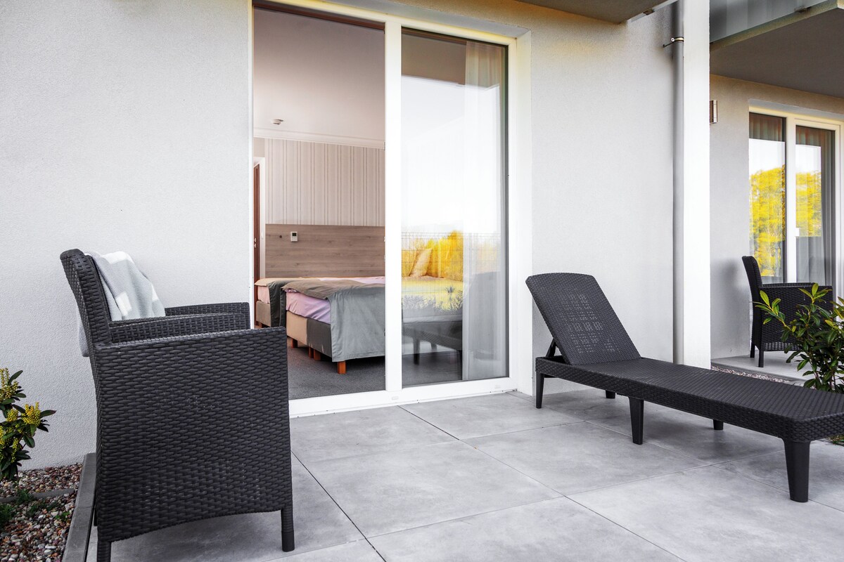 A balcony with two wicker chairs and a lounge chair, a bed with a yellow bedspread, and a sliding glass door leading to an indoor room.