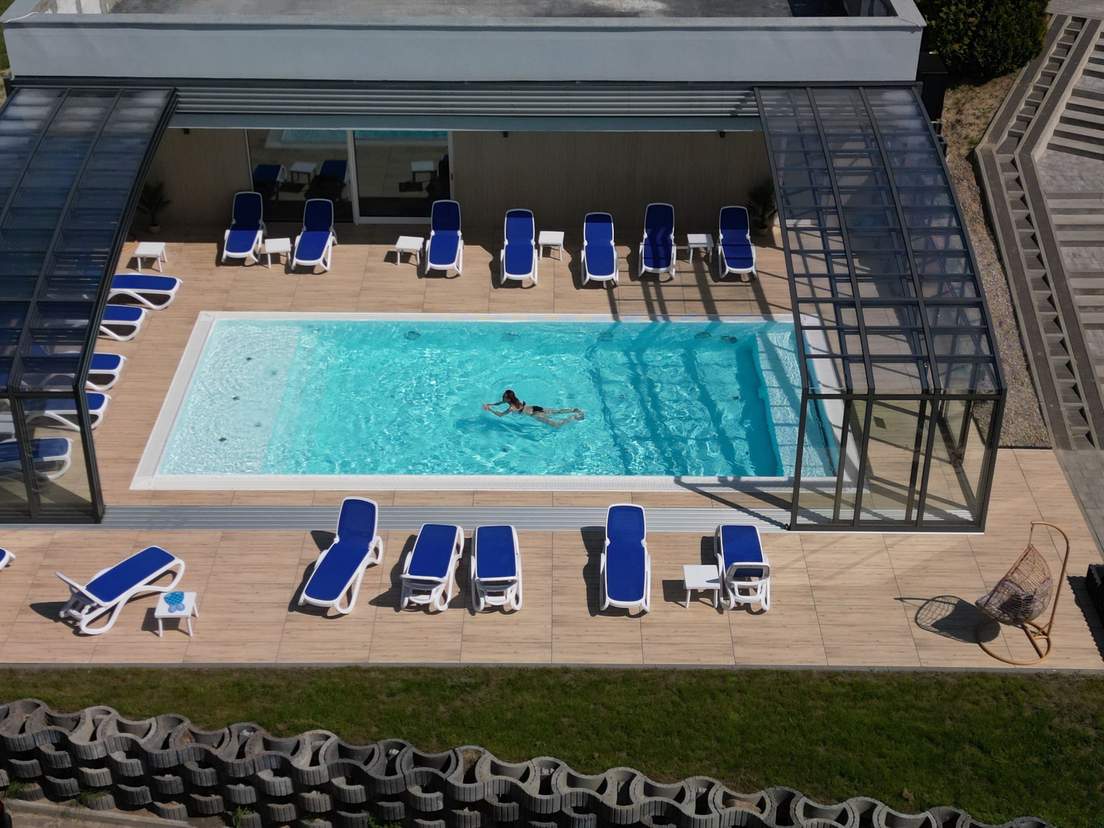 Une piscine sur le toit, où une personne nage, entourée de chaises longues et protégée par une paroi en verre.