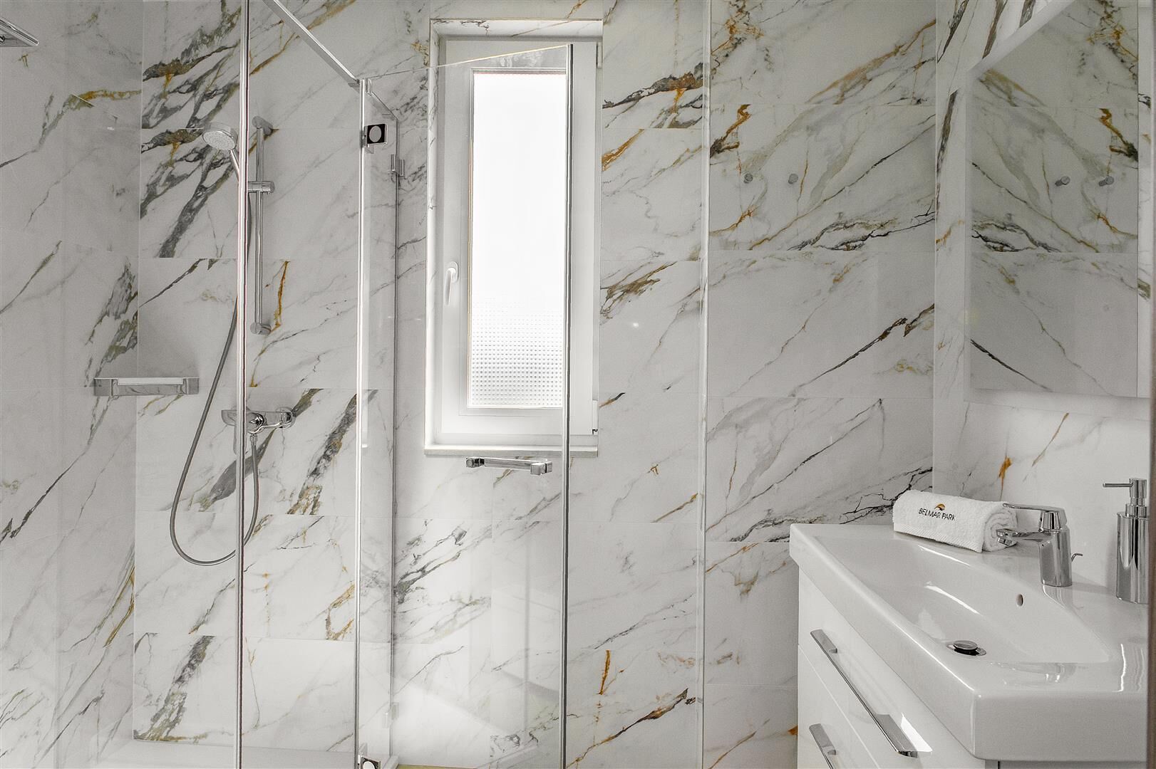 A bathroom with a glass-enclosed shower, a white sink, and marble walls.