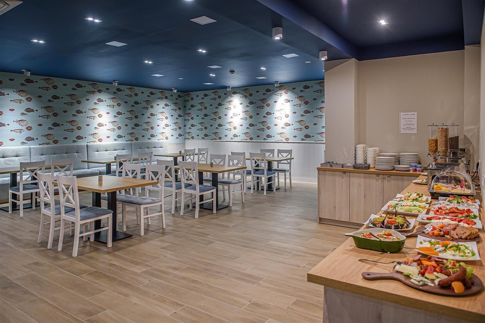 Een restaurant met een blauw plafond, witte stoelen en een buffetgedeelte met diverse gerechten.