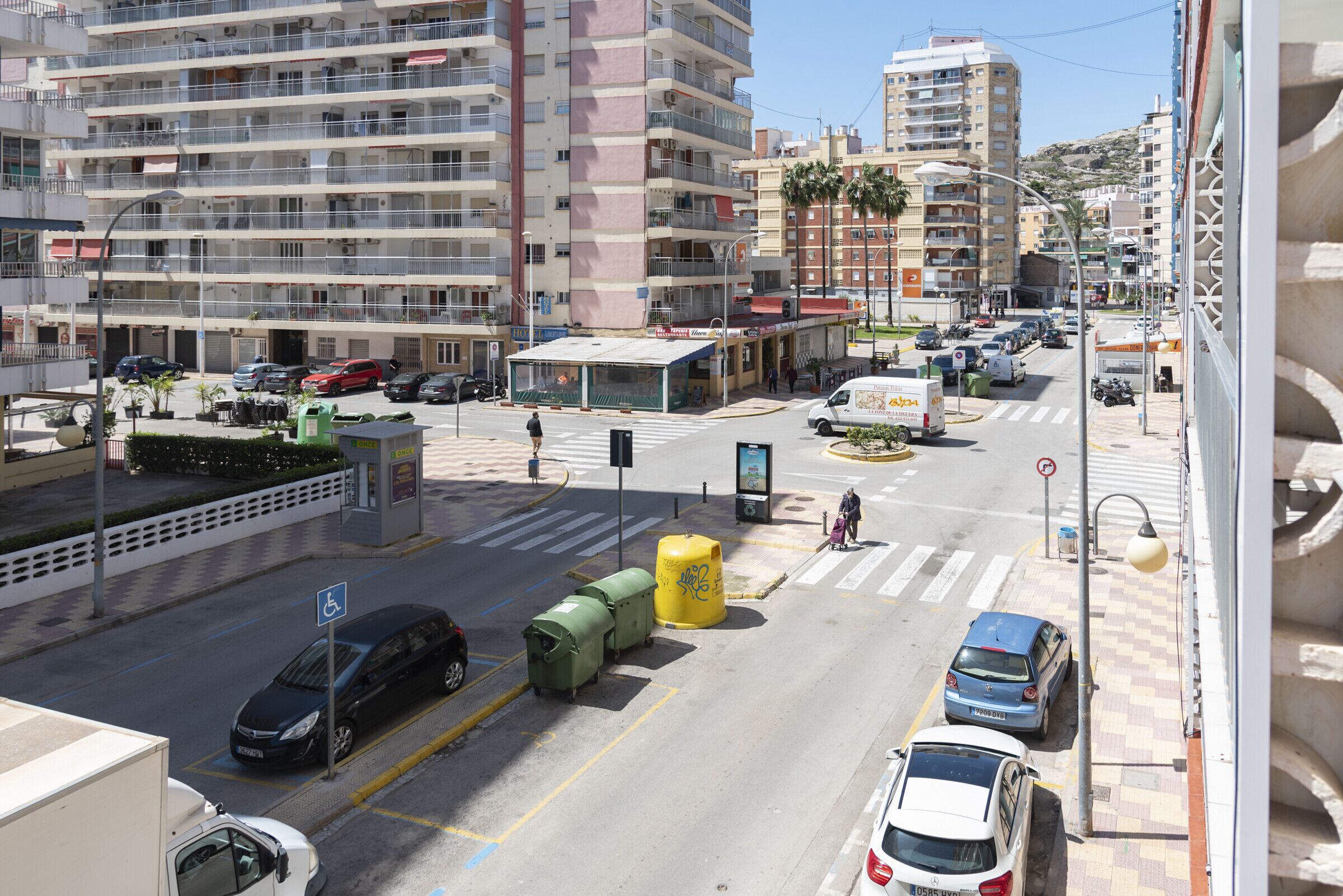 Une rue animée en milieu urbain, bordée d’immeubles à plusieurs étages, avec un passage piéton et des voitures garées.