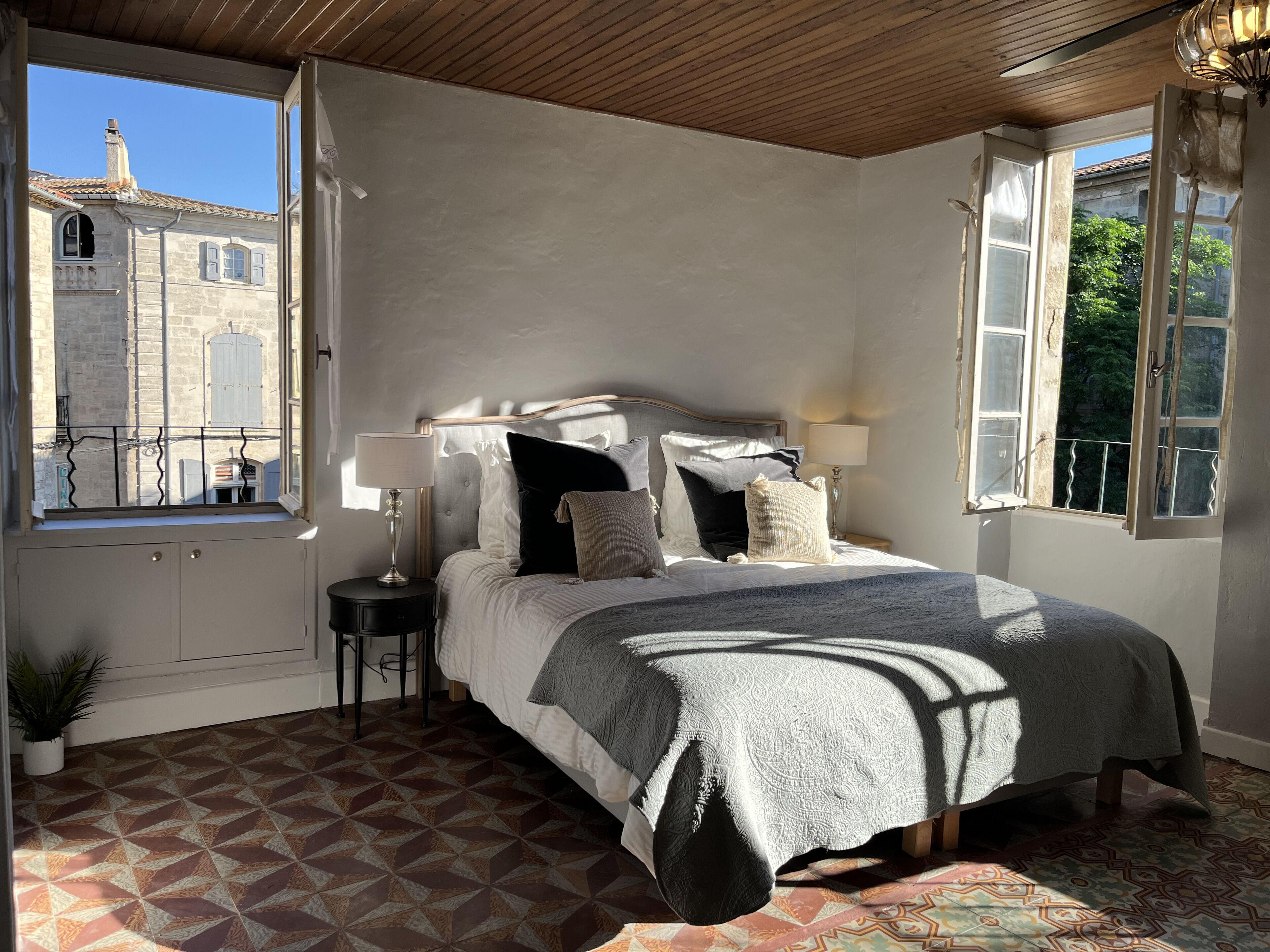 Une chambre à coucher avec un grand lit, deux tables de chevet, une fenêtre donnant sur un immeuble et un sol à motifs.