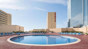 A swimming pool with lounge chairs, a tall building labeled "SOMOS BOAT" in the background, and modern high-rise buildings. - Beachfront Double Queen Hotel CozySuite (Atlantic City)