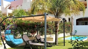 A patio with a swing chair, two lounge chairs, a pool, and a covered seating area.