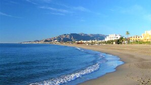 A beach with a clear view of the ocean and a mountain in the background. There are buildings along the shore.