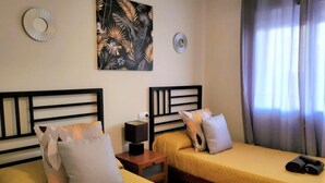 A hotel room with two beds, a nightstand with a lamp, a wall clock, and a tropical-themed painting.