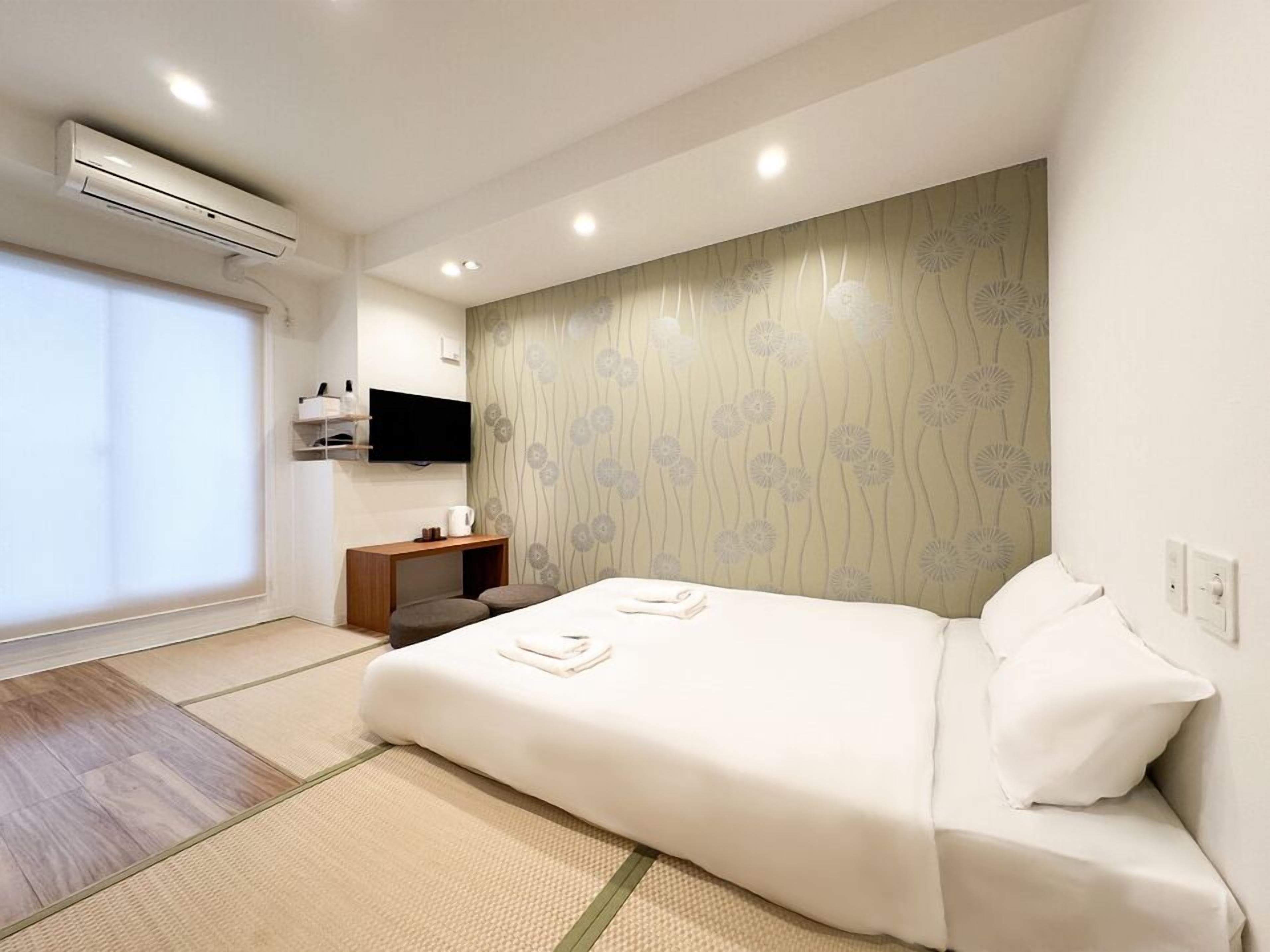 A modern hotel room with a bed, a small wooden side table, a mounted TV, and a large window.