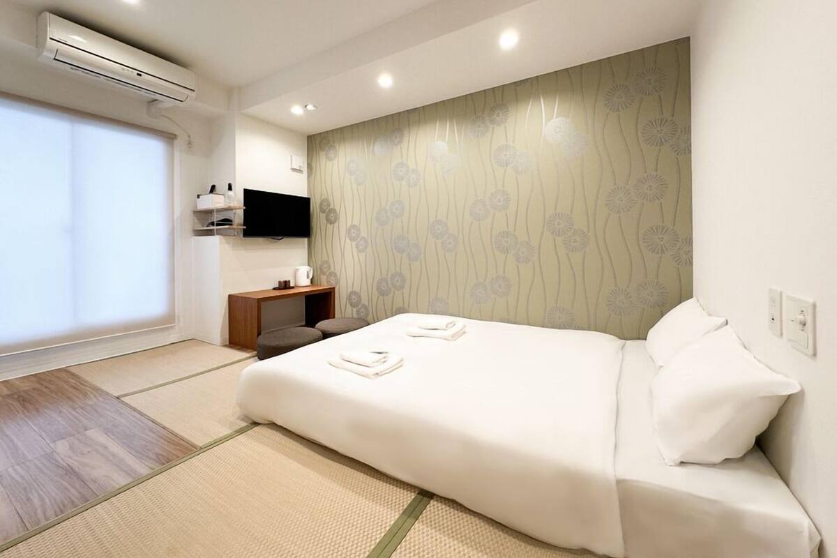 A modern hotel room with a bed, a small wooden side table, a mounted TV, and a large window.