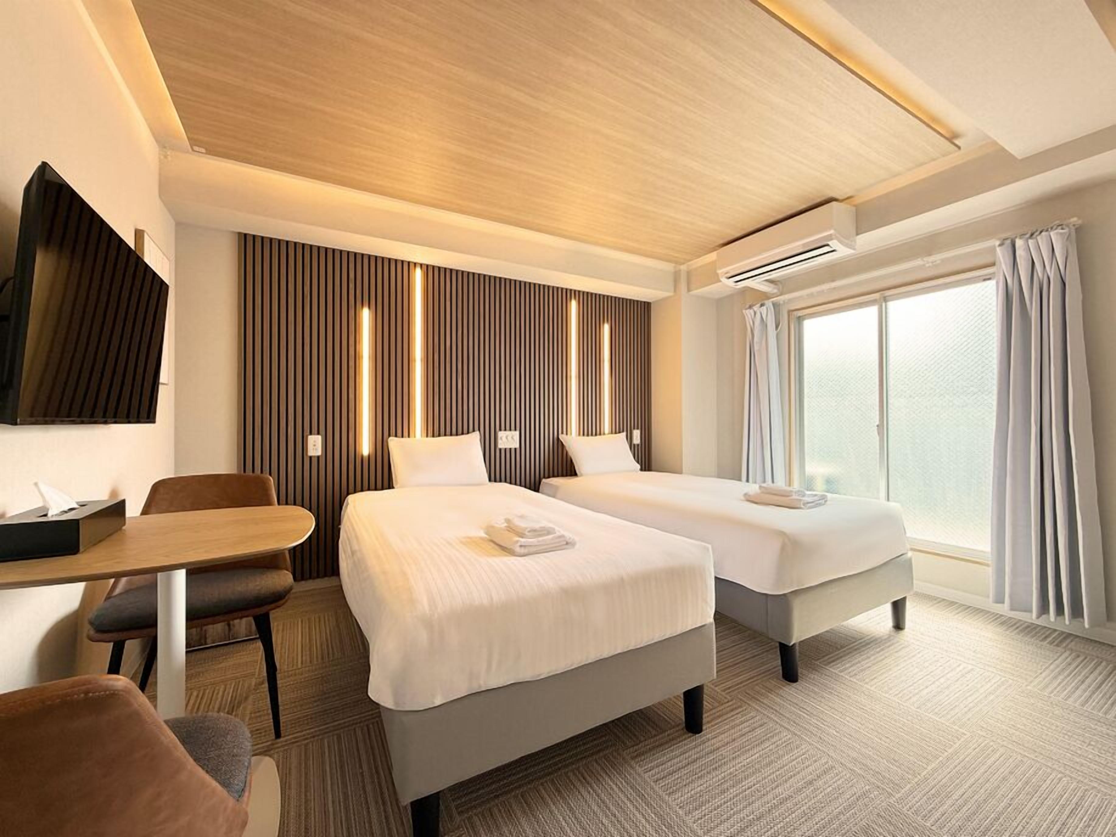 A hotel room with two beds, a desk, a chair, a TV, and a large window.