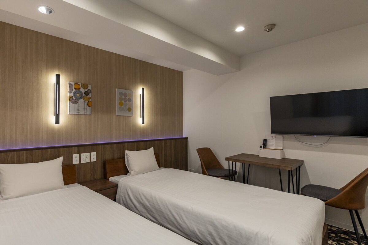 A hotel room with two beds, a desk, a chair, a flat-screen TV, and wall-mounted artwork.