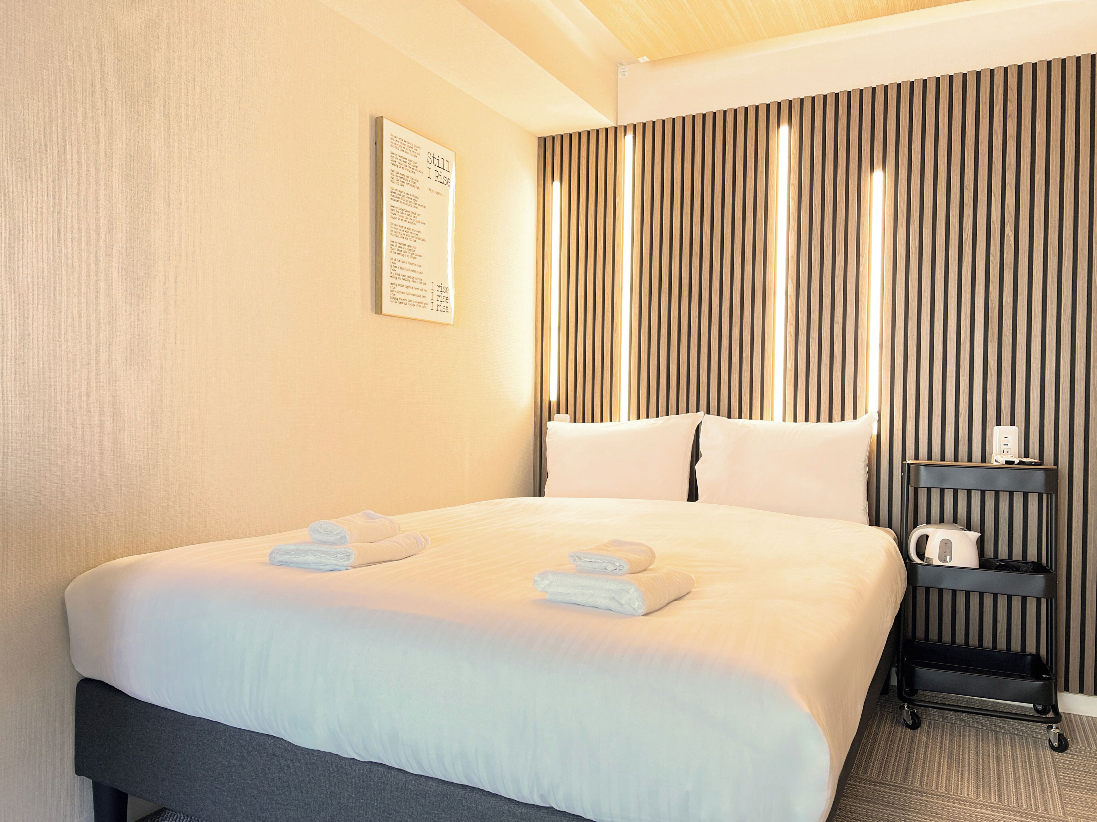 A hotel room with a large bed, a bedside table with a kettle, and a wall-mounted sign.