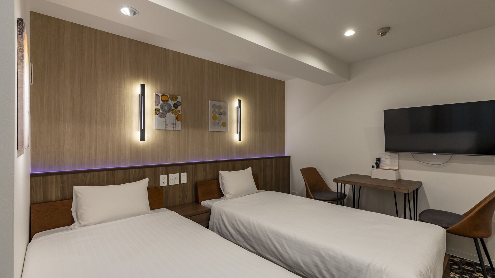 A hotel room with two beds, a desk, a chair, a flat-screen TV, and wall-mounted artwork.