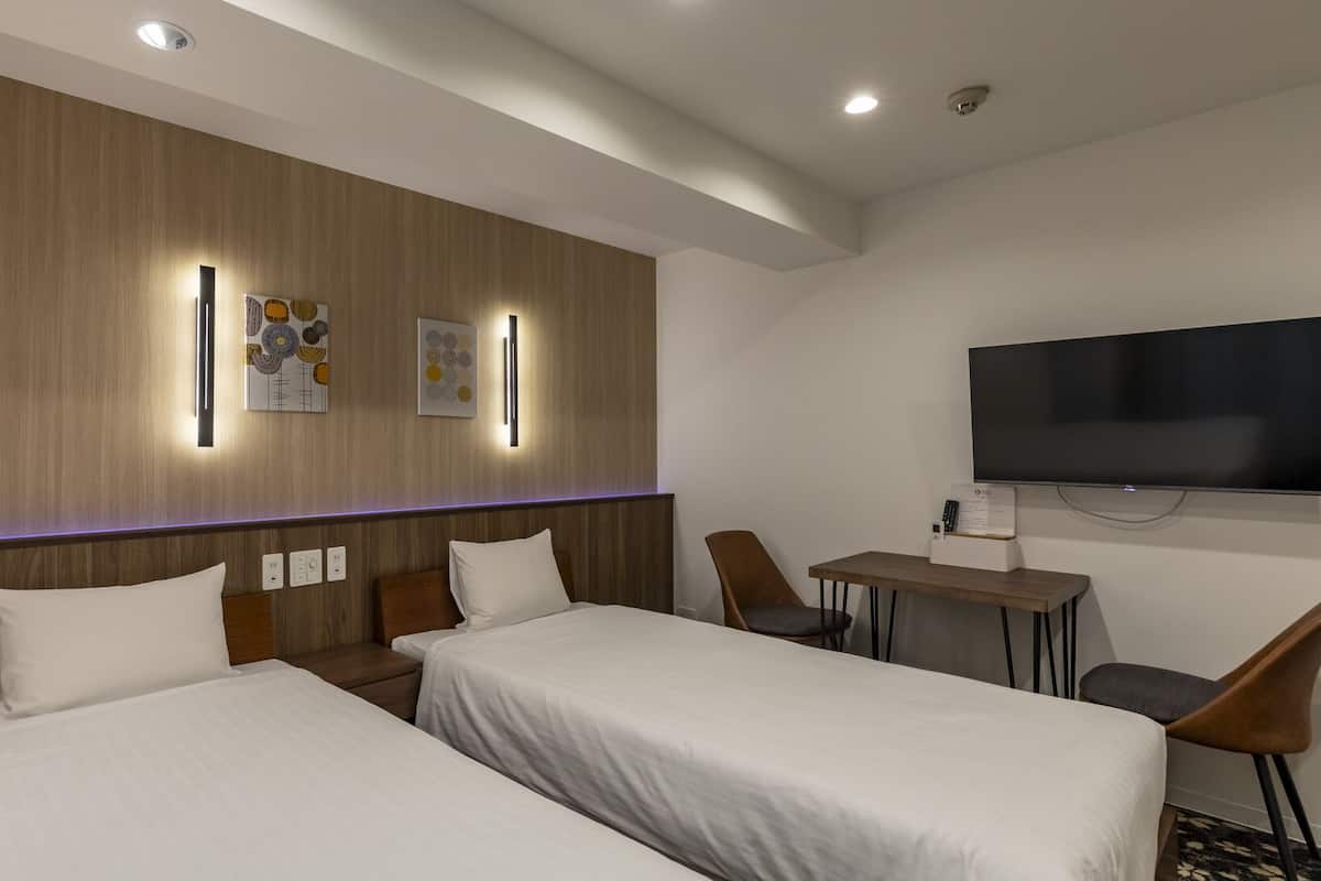 A hotel room with two beds, a desk, a chair, a flat-screen TV, and wall-mounted artwork.