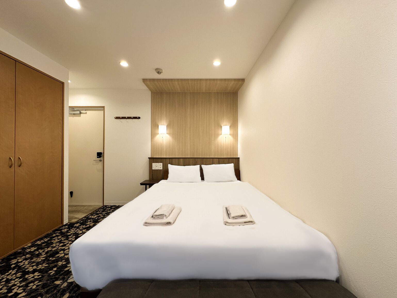 A hotel room with a bed, two bedside lamps, a wooden headboard, and a carpeted floor.