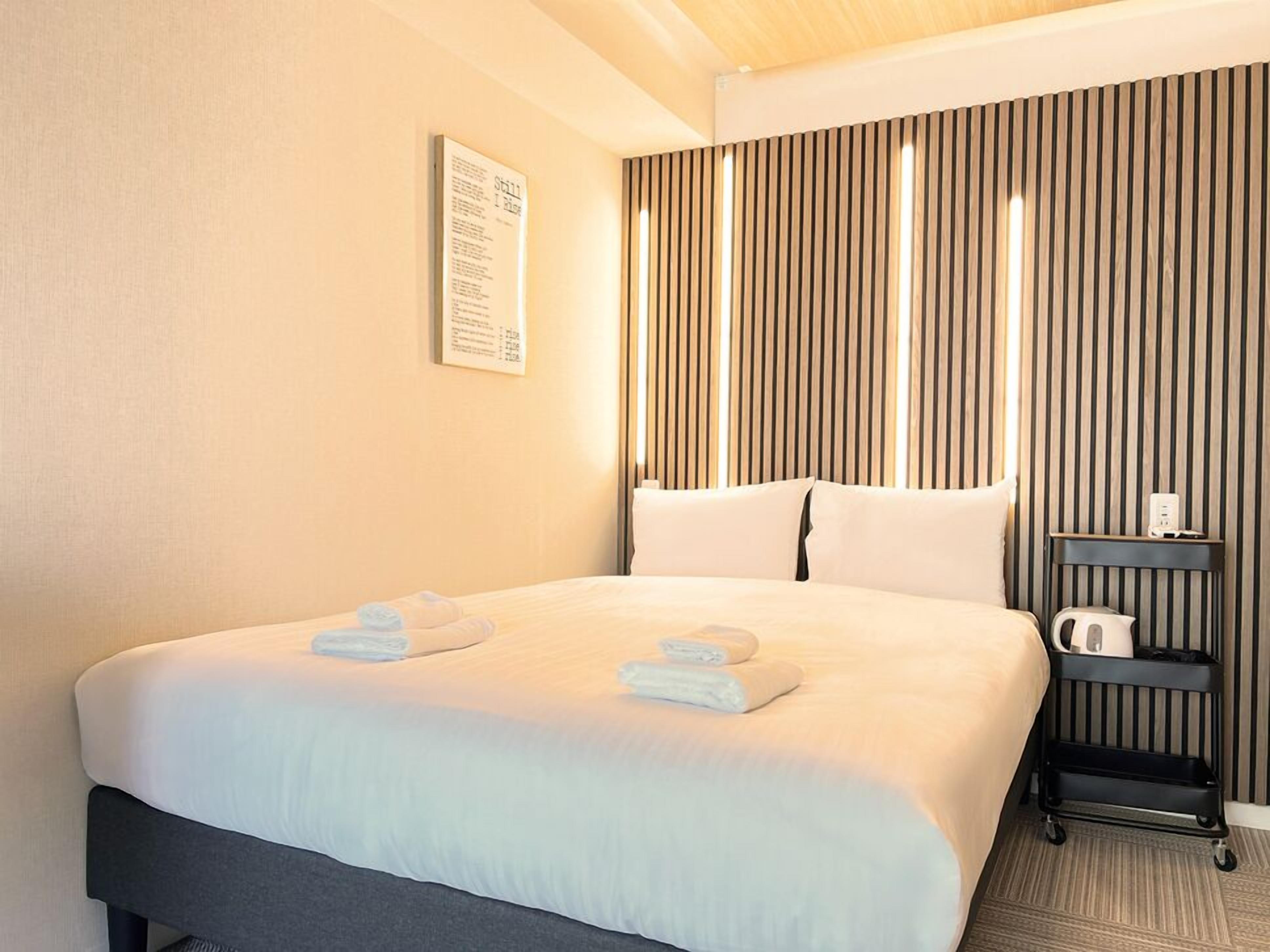 A hotel room with a bed, pillows, a nightstand with a kettle, and a wall-mounted sign.