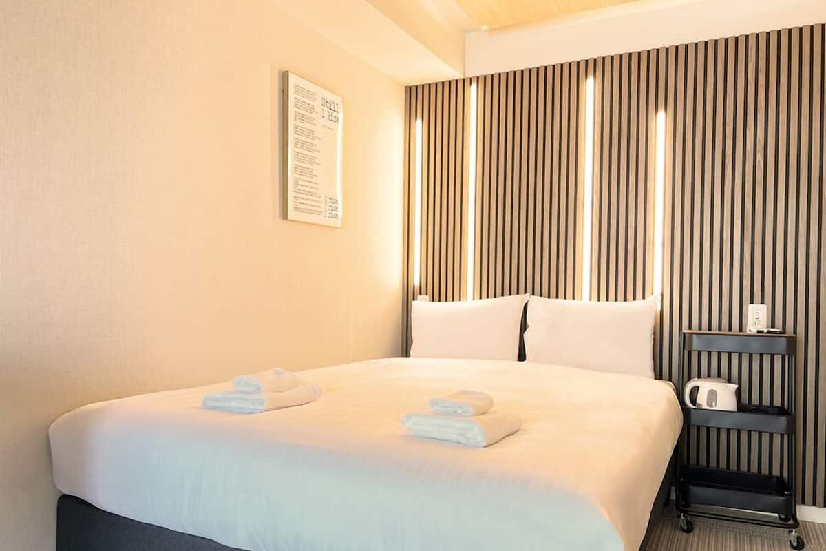 A hotel room with a bed, pillows, a nightstand with a kettle, and a wall-mounted sign.