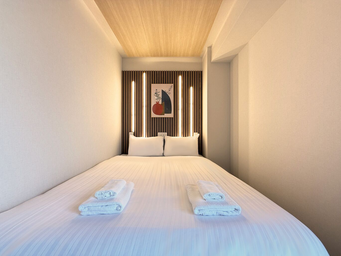 A modern hotel room with a large bed, two pillows, a headboard with vertical slats, and a framed artwork on the wall.