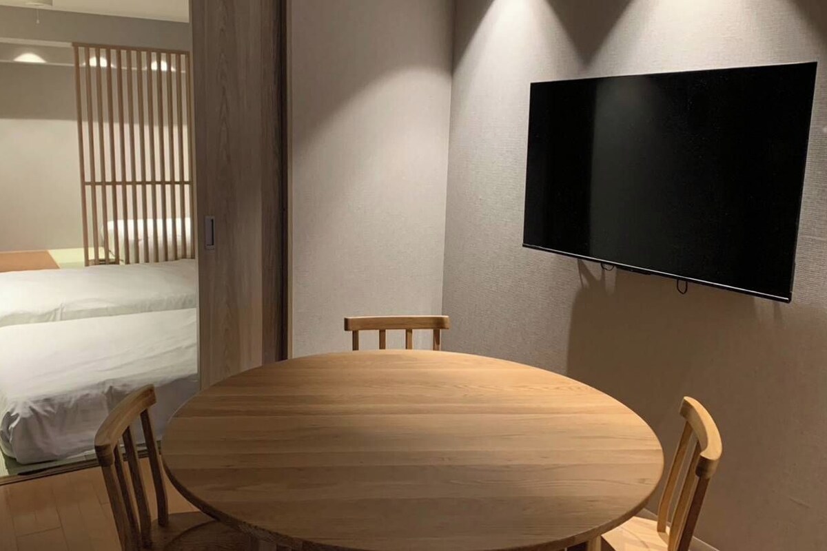 A room with a bed, a round wooden table, two chairs, a TV mounted on the wall, and a window with curtains.