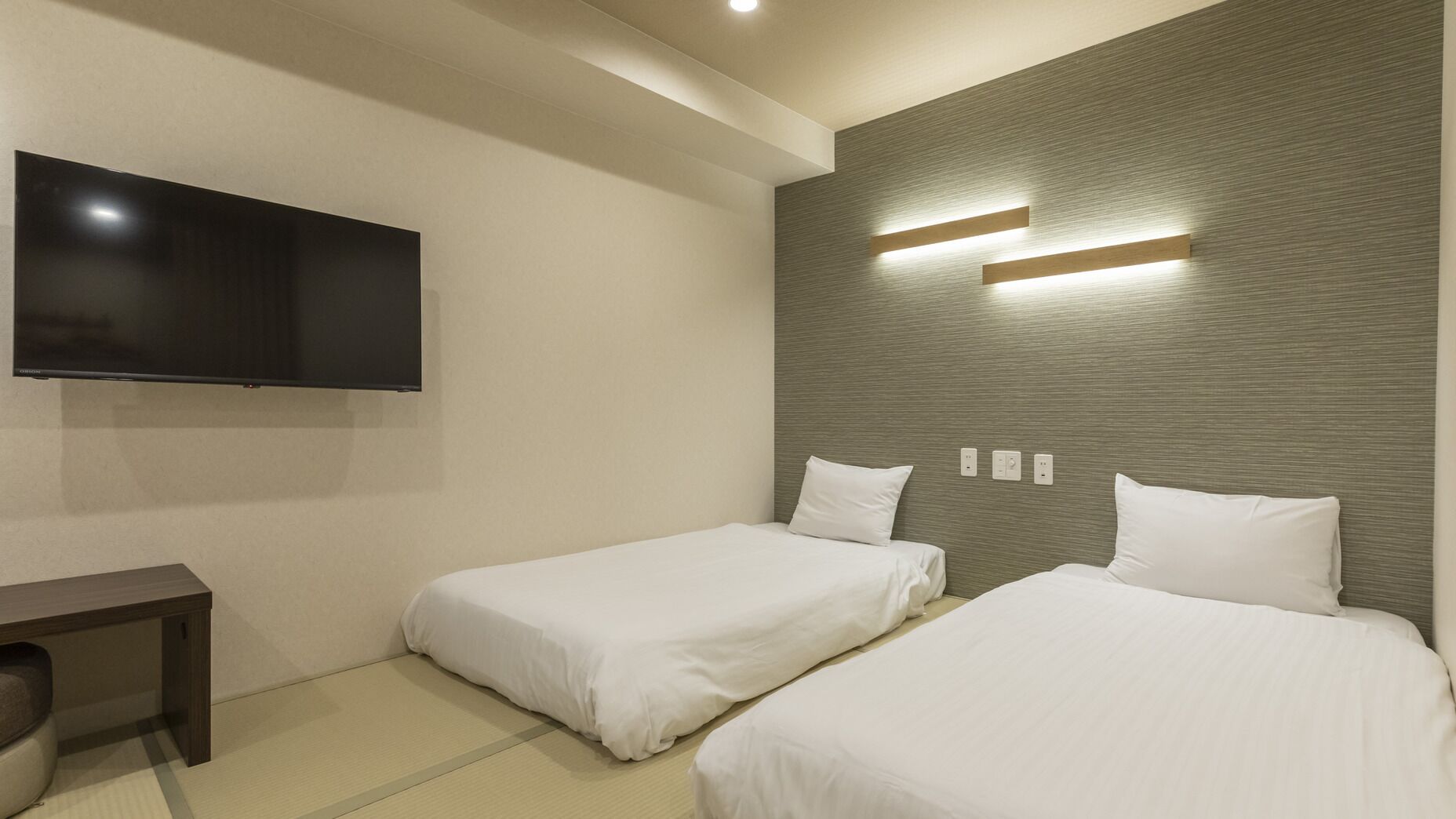 A hotel room with two beds, a flat-screen TV, a desk, and a wall-mounted light fixture.