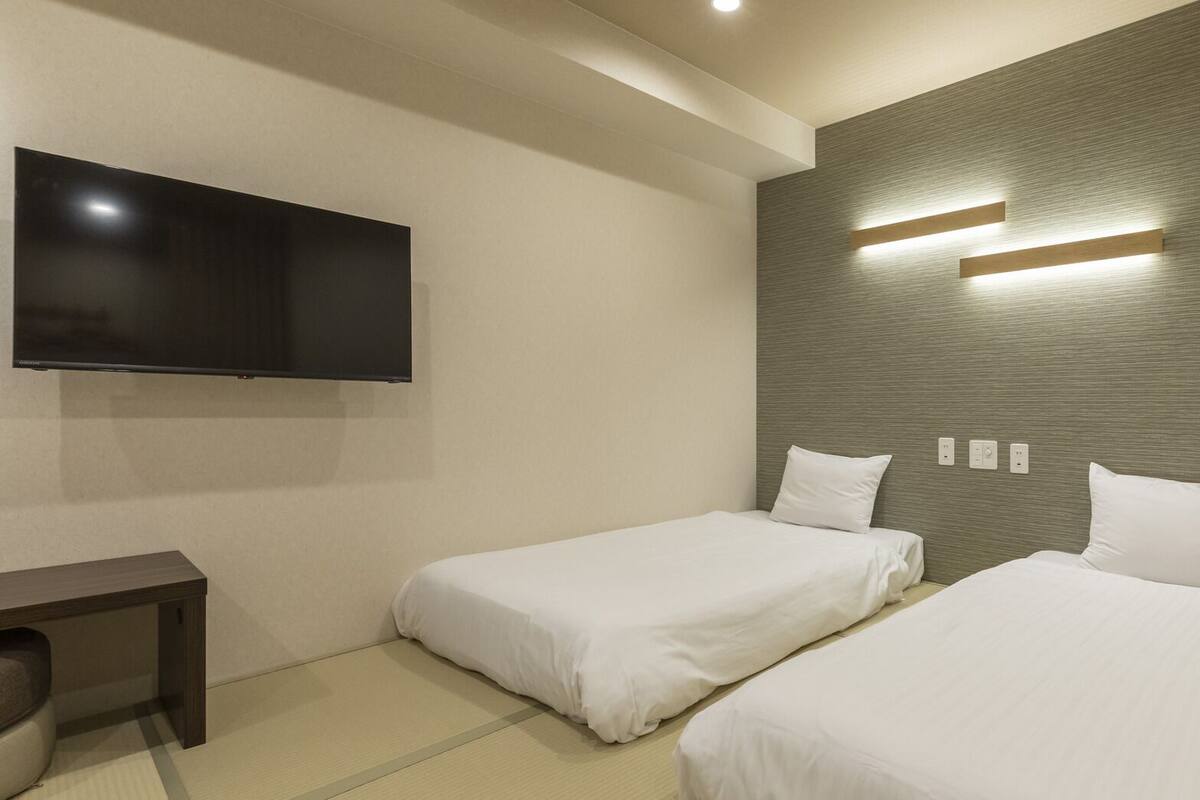 A hotel room with two beds, a flat-screen TV, a desk, and a wall-mounted light fixture.