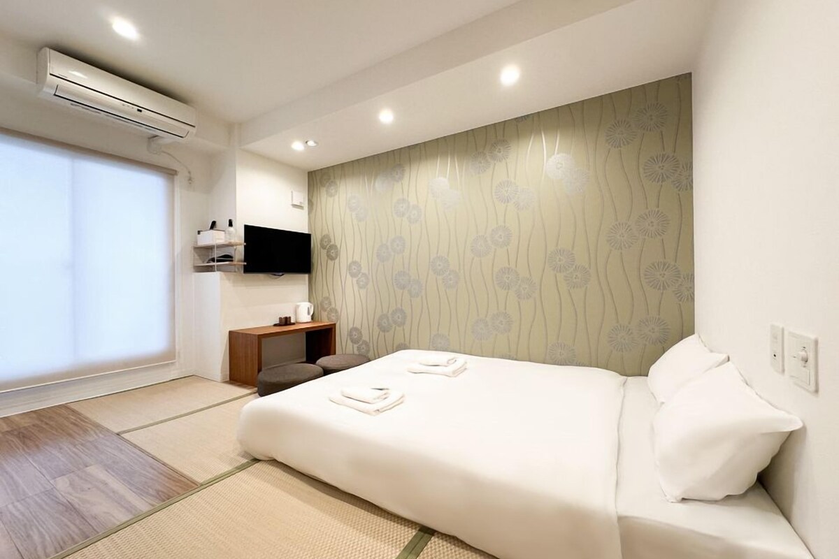 A modern hotel room with a bed, a small wooden side table, a mounted TV, and a large window.