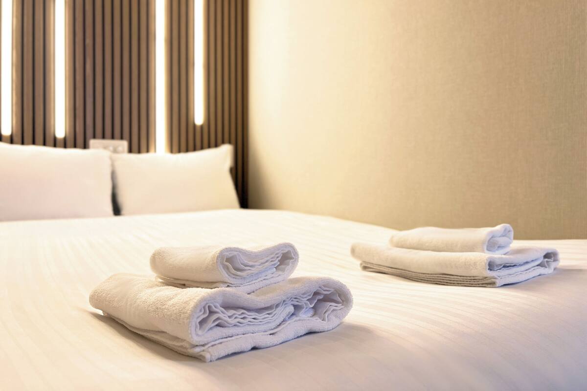 A neatly made bed with white linens and folded towels, a wooden headboard, and a neutral-colored wall.