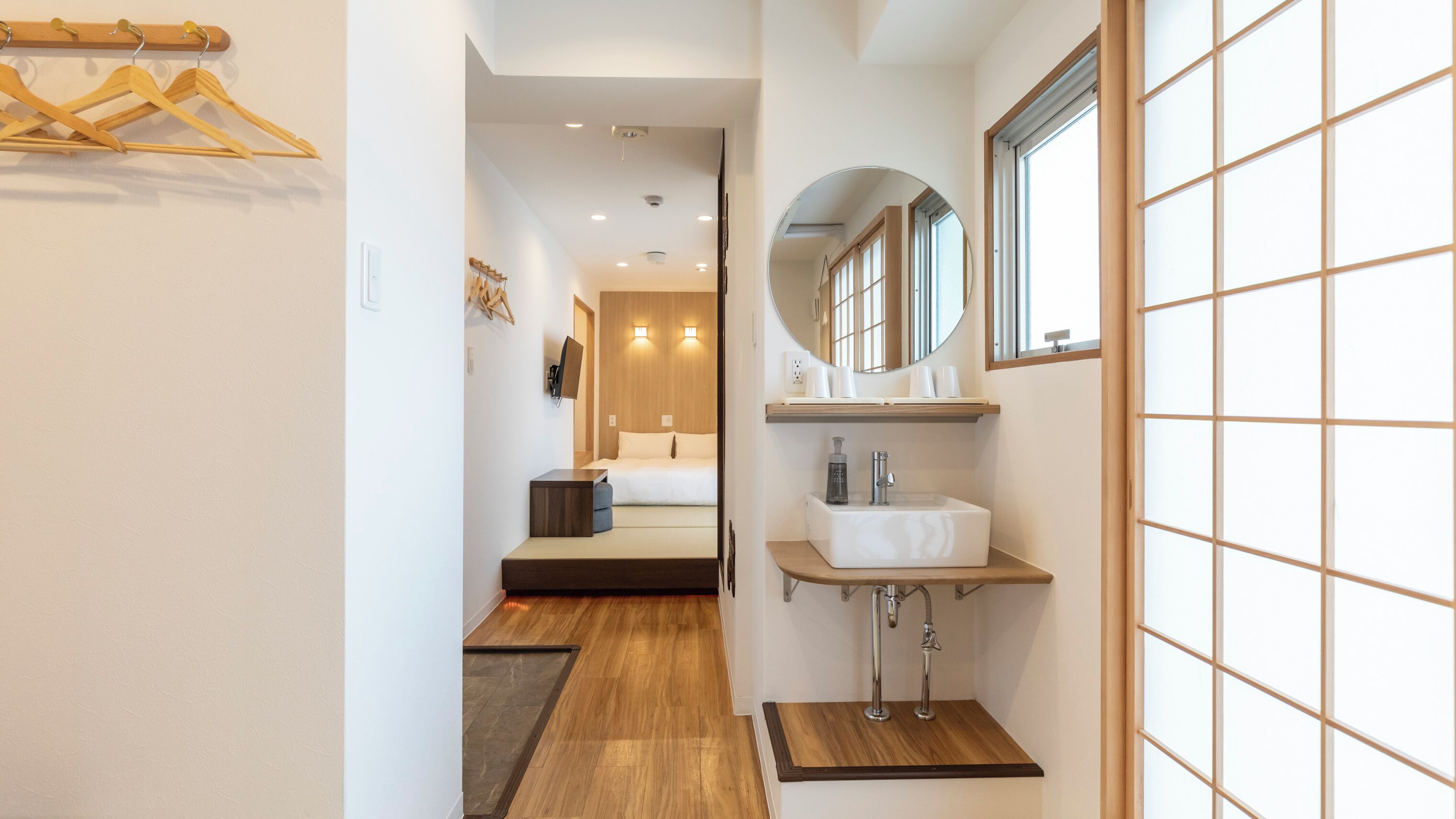 A hotel hallway with a bed, a bathroom with a sink and mirror, and wooden flooring.